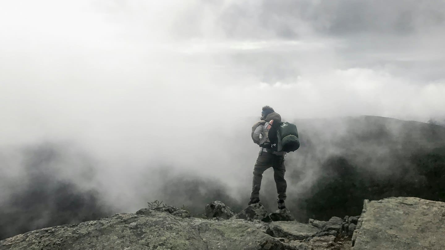 Backpacker standing on a rocky mountain peak overlooking misty valleys.