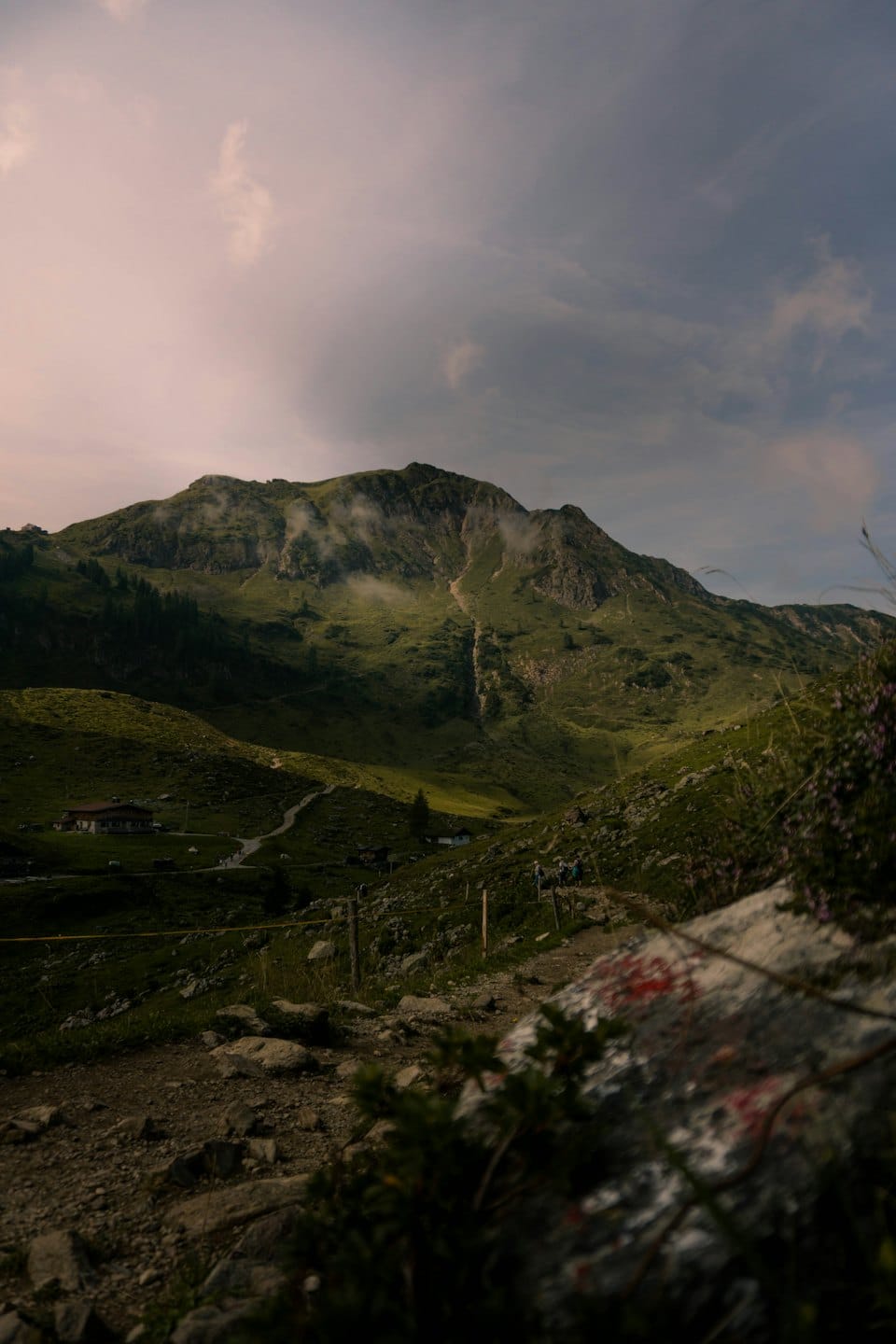 Hikers walk along a mountain path toward distant small buildings.